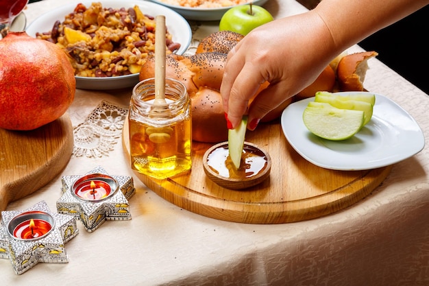 De hand van een vrouw doopt een stuk appel in honing ter ere van de viering van Rosj Hasjana in de buurt van honing en challah en traditioneel eten