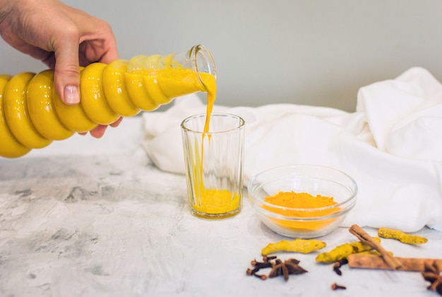 De hand van een vrouw giet een kurkuma latte met melk en kaneel uit een fles in een glas. een elixer van gezondheid en kracht. een traditionele gezonde indiase detoxdrank. grijze achtergrond