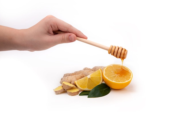 De hand van een vrouw houdt een houten honinglepel vast en giet honing over een citroen. Geïsoleerde stukjes gember en citroen met honing.