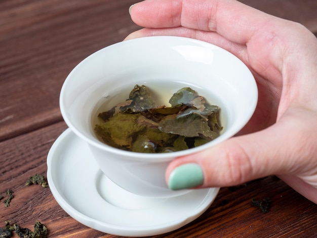 De hand van een vrouw houdt een kom van wit porselein met thee vast. Houten achtergrond. De geopende bladeren van grootbladige thee. Chinese thee. theekransje
