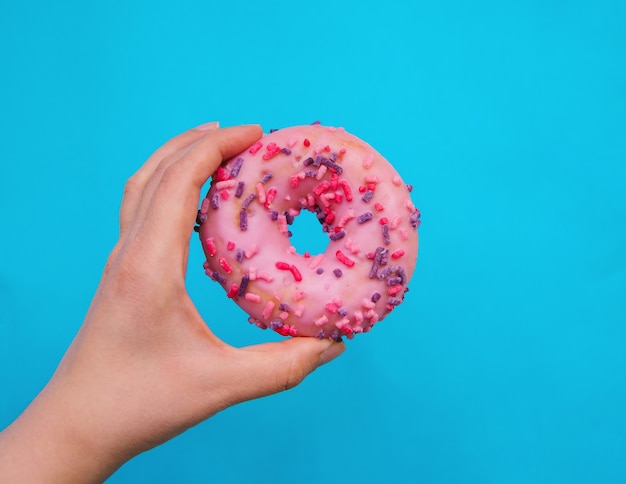 De hand van een vrouw houdt een roze donut op een blauwe achtergrond. Zoetwaren. Schadelijk fastfood. De donut is bedekt met glazuur en versierd met een eetbaar decor.