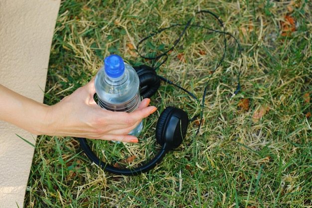 De hand van een vrouw neemt een fles water naast de koptelefoon op een grasveld Hydratatieconcept