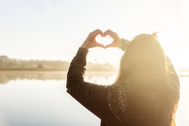 De handen die van de vrouw een hartvorm vormen op ochtendzonsopgang.