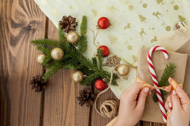 De handen die van de vrouw Kerstmisgift verpakken, sluiten omhoog. Onbereide kerstcadeautjes op houten met decorelementen en items, bovenaanzicht. Kerstmis of Nieuwjaar DIY-verpakking.