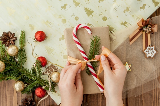 De handen die van de vrouw Kerstmisgift verpakken, sluiten omhoog. Onbereide kerstcadeautjes op houten met decorelementen en items, bovenaanzicht. Kerstmis of Nieuwjaar DIY-verpakking.