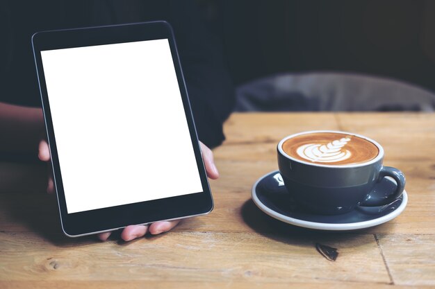 De handen die van de vrouw zwarte tablet met het witte lege scherm en de koffiekop op houten lijst tonen