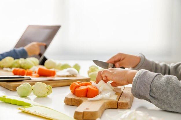 De handen die van een meisje voedsel in de keuken voorbereiden terwijl haar broer de tablet bekijkt