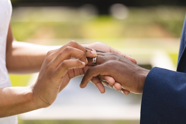 De handen van de Afro-Amerikaanse bruidegom en bruid plaatsen een ring op de vinger bij de huwelijksceremonie in de zonnige tuin