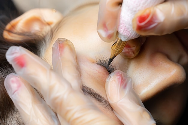 De handen van de meester van permanente make-up die de tatoeage van het ooglid van het model uitvoert, zijn over haar gebogen