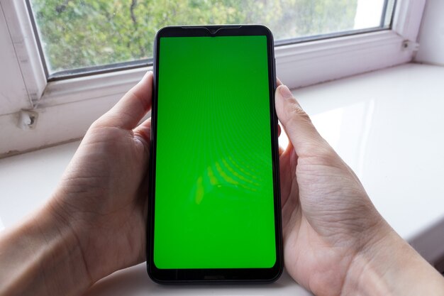 De handen van de vrouw houden de smartphone in een verticale positie met een groen scherm op een raam, chroma key, mock-up.