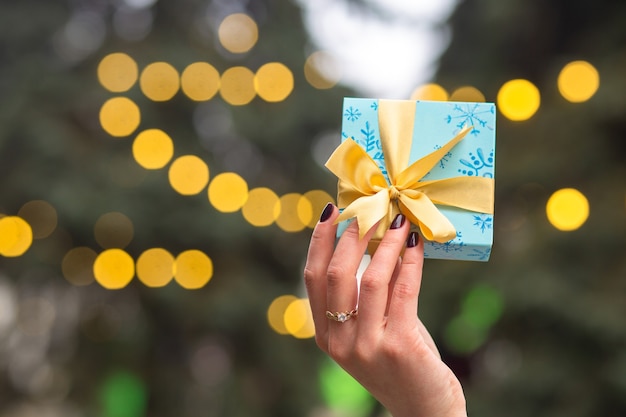 De handen van de vrouw houden een kleine geschenkdoos vast op de achtergrond van de kerstboom met vage lichten. Lege ruimte