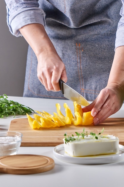 De handen van de vrouw snijden biologische paprika's op een houten bord op een grijze tafel. Salade koken.