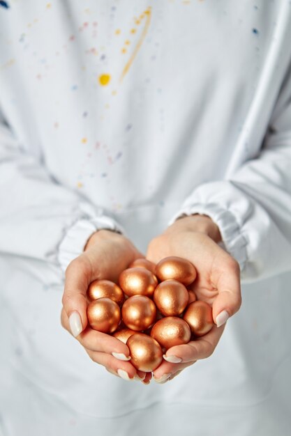 De handen van een jong meisje houden gouden beschilderde eieren vast op de achtergrond van een schort met verfbrikjes