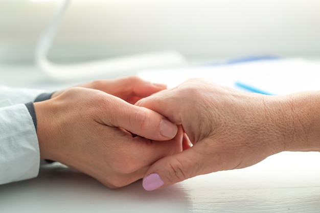 De handen van een jonge dokter houden de handen van een oudere geduldige vrouw vast als een teken van steun.