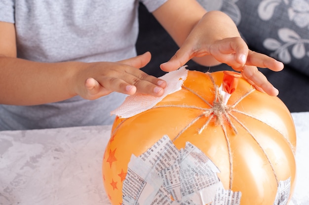 Foto de handen van een kind lijmen servetten aan een ballon om een pompoen van papier-maché te maken voor halloween-decor.
