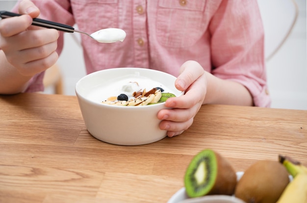 De handen van een meisje in een roze shirt houden een kom yoghurtgranola en vers fruit op een houten tafel Het concept van gezond eten