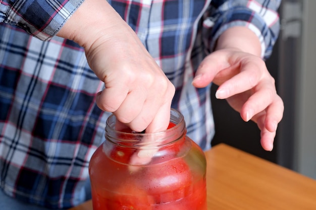 De handen van een oudere vrouw halen heerlijke zelfgemaakte gepekelde tomaten uit een pot in de keuken aan een bruine tafel zonder gezichtsclose-up