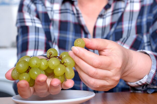 Foto de handen van een oudere vrouw houden een klein penseeltje witte pitloze druiven vast en tonen ze in de keuken aan een bruine tafel zonder gezichtsclose-up