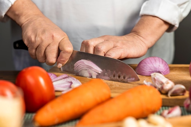 De handen van een volwassen vrouw die ui snijdt met een keukenmes op een houten bord voor de bereiding van lasagne