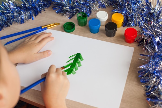 De handen van het kind houden een penseel vast en halen gouache uit een pot Hobby's en amusement voor kinderen Close-uptekening van een kerstboom