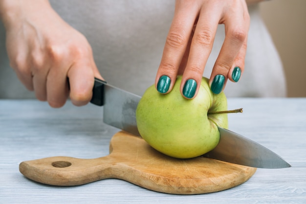 De handen van vrouwen met een heldere manicure snijden groene appel met een keukenmes