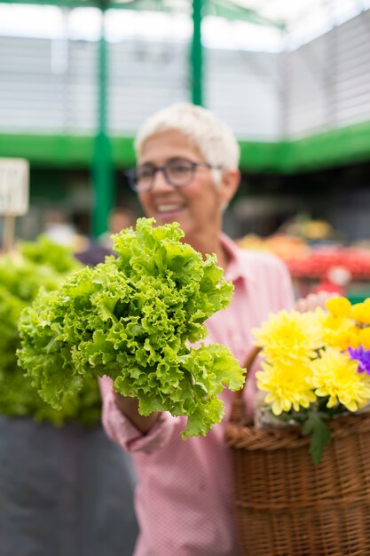 De hogere vrouw houdt mand met bloemen en koopt sla op markt