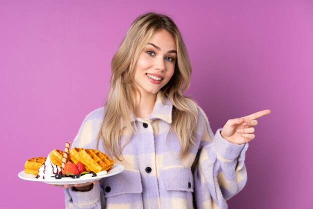 Foto de holdingswafels van de tienervrouw die op purpere muur worden geïsoleerd die vinger aan zij richten en gelukkig