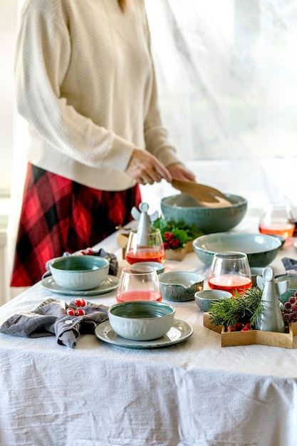 De instelling van de rustieke kersttafel met leeg ambachtelijk keramisch serviesgoed, borden en kommen, kerst engel decoraties op wit tafellaken voor het raam. Vrouw houdt een slakom