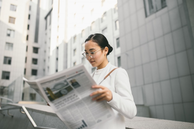 Foto de jonge aantrekkelijke aziatische vrouwelijke bankier of de accountant in glazen leest krant buiten een modern bureaucentrum