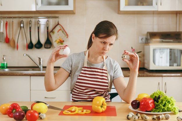 De jonge aantrekkelijke peinzende vrouw in een schort kiest tussen kip en kwarteleitjes in de keuken. Dieet concept. Gezonde levensstijl. Thuis koken. Eten koken.