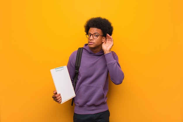 De jonge Afrikaanse Amerikaanse studentenmens die een klembord houden probeert aan het luisteren van een roddel