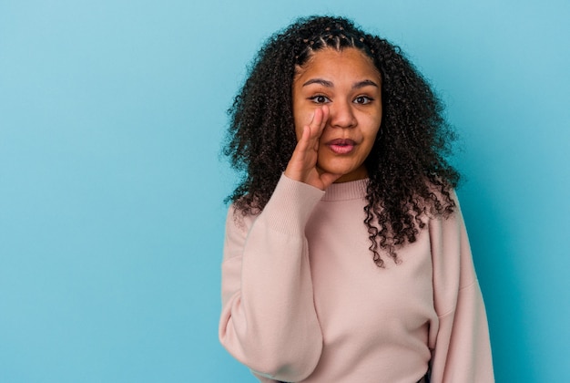 De jonge afrikaanse amerikaanse vrouw die op blauwe muur wordt geïsoleerd, zegt een geheim heet remmend nieuws en kijkt opzij