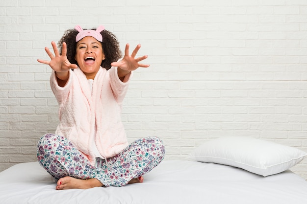 De jonge afrikaanse amerikaanse vrouw in het bed dat pijama draagt voelt zeker het geven van een knuffel aan de camera.