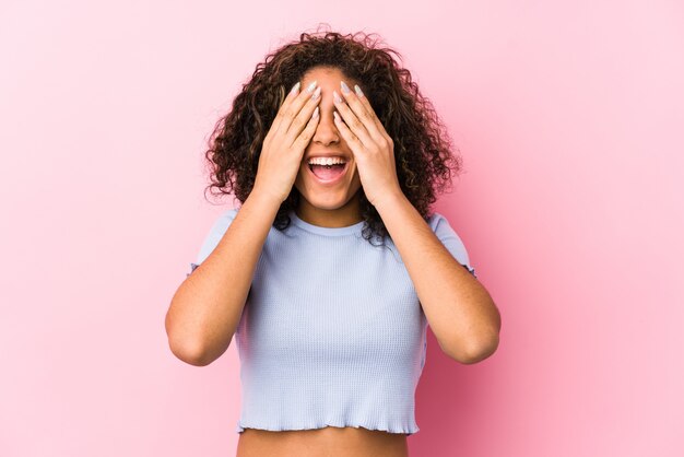 De jonge Afrikaanse Amerikaanse vrouw tegen een roze muur behandelt ogen met handen, glimlacht breed wachtend op een verrassing.