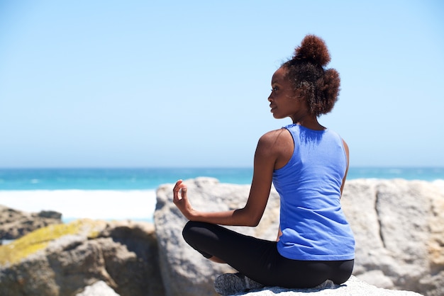 De jonge Afrikaanse vrouwenzitting bij strand in yoga stelt