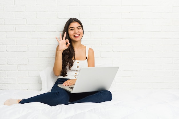 De jonge Arabische vrouw die met haar laptop aan het bed werkt knipoogt een oog en houdt een ok gebaar met hand.