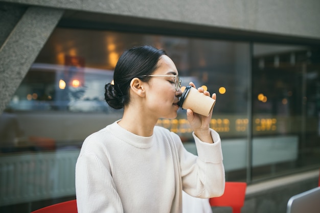 De jonge Aziatische bedrijfsvrouw in glazen zit buiten een koffiewinkel, drinkend koffie