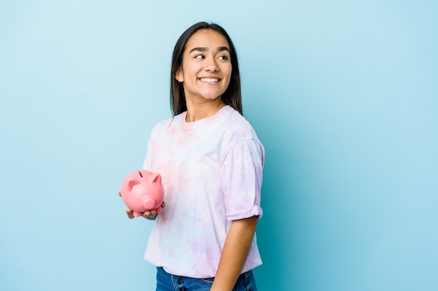 De jonge Aziatische vrouw die een roze bank over geïsoleerde muur houdt kijkt glimlachend, vrolijk en aangenaam opzij.