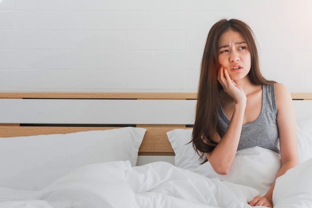 De jonge Aziatische vrouw voelt tandpijn en ongemak op bed in witte slaapkamerochtend.