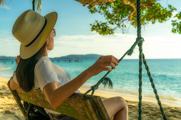 De jonge Aziatische vrouw zit en ontspant op schommeling bij kust op de zomervakantie. Zomerse vibes. Vrouwenreis alleen op vakantie. Backpacker bij tropisch paradijsstrand.