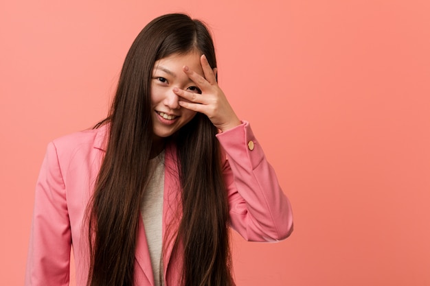 De jonge bedrijfs Chinese vrouw die roze kostuum draagt knipoogt naar de camera door vingers, beschaamd behandelend gezicht.