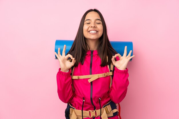 De jonge bergbeklimmervrouw met een grote rugzak over geïsoleerde roze muur in zen stelt