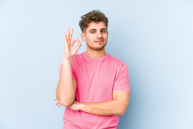 De jonge blonde krullende geïsoleerde haar Kaukasische mens knipoogt een oog en houdt een ok gebaar met hand.