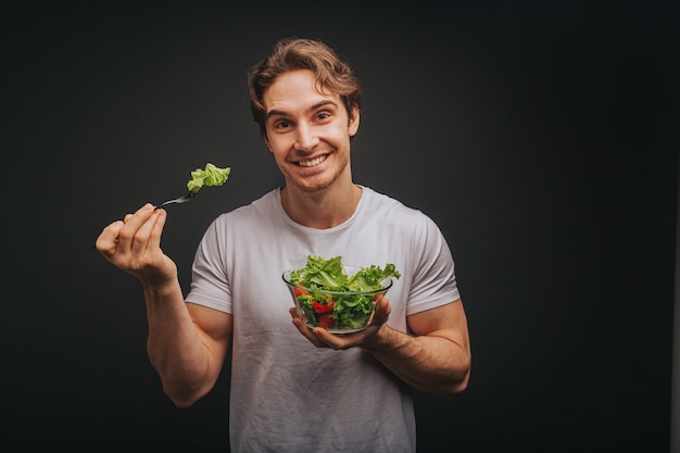 De jonge blonde mens in wit t-shirt houdt gelukkig plaat van salade