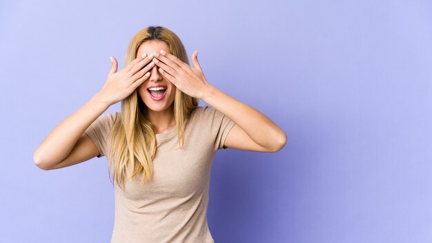 Foto de jonge blonde vrouw behandelt ogen met handen, in grote lijnen glimlacht wachtend op een verrassing