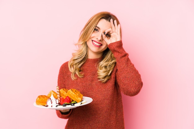 De jonge blondevrouw die een wafeldessert eten isoleerde opgewekt houdend ok gebaar op oog