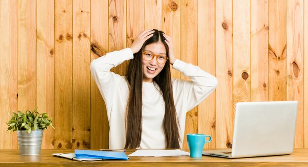 De jonge Chinese vrouw die op haar bureau bestudeert lacht vreugdevol houdend handen op hoofd.