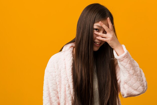 De jonge Chinese vrouw in pyjama knipoogt door door vingers, beschaamd behandelend gezicht.