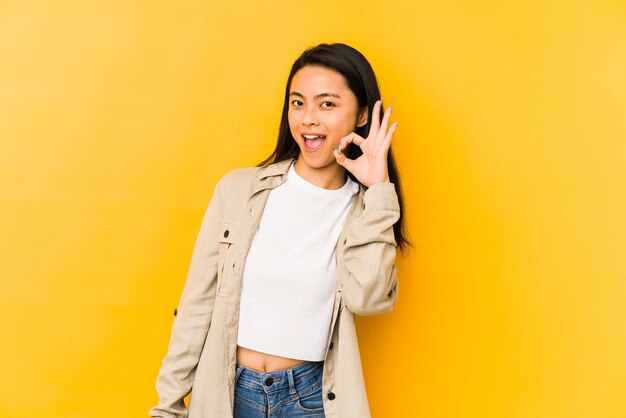 De jonge chinese vrouw op een gele muur knipoogt een oog en houdt een ok gebaar met hand.