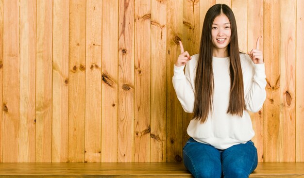 De jonge Chinese vrouwenzitting op een houten plaats wijst op met beide voorvingers die omhoog een lege ruimte tonen.